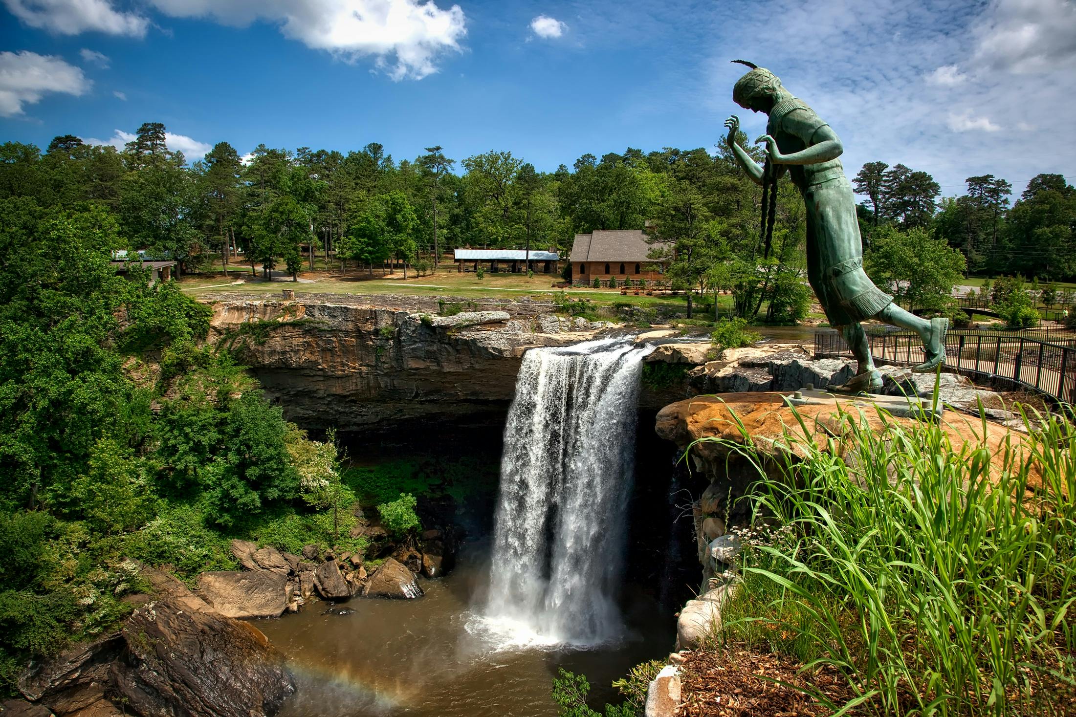 A breathtaking view of Noccalula Falls with a historic statue amidst lush greenery under a bright sky.